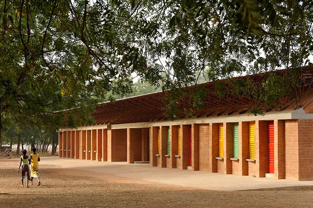 francis-kere-vencedor-pritzker-2022-erik-jan-owerkerk-escola-gando-fachada Imagem mostra árvore grande e fachada da escola em Gando, construída com tijolos aparentes e janelas coloridas em amarelo, verde e vermelho.
