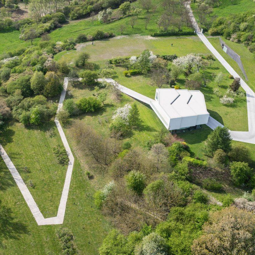 quem-disse-concreto-precisa-ser-cinza-olo-studio-casa-fachada-concreto-branco Foto aérea mostra estrada de concreto branco em meio à paisagem verde, com uma casa inteira na cor branca.