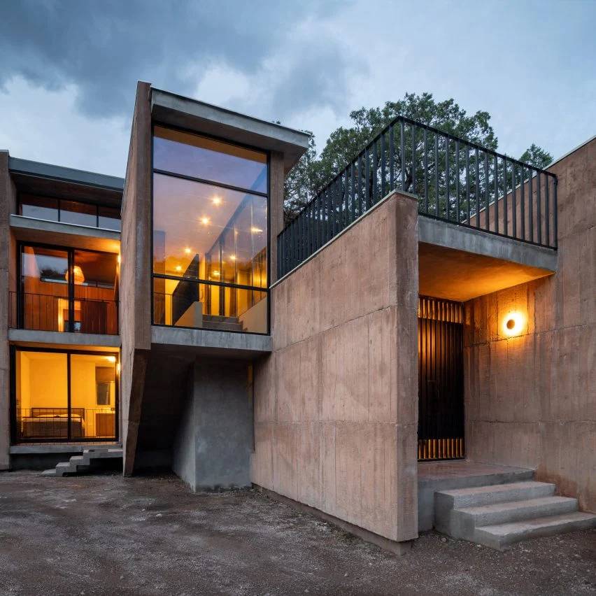 Casa com fachada em linhas retas e tom de terra natural, aberturas em vidro e iluminação amarelada, ao entardecer.