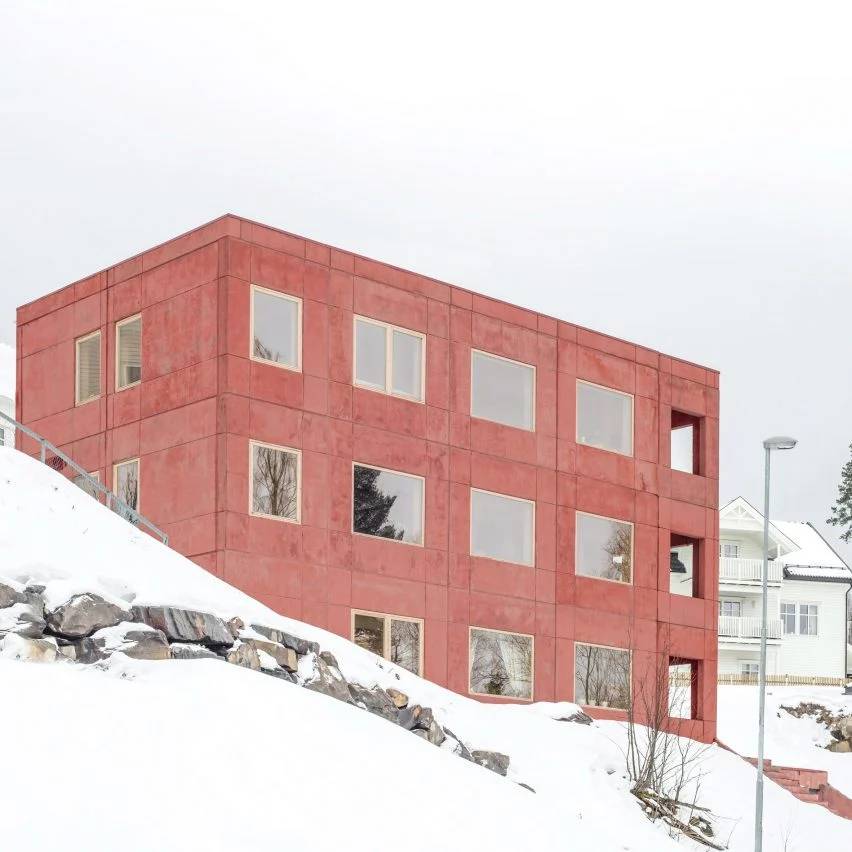 quem-disse-concreto-precisa-ser-cinza-sanden-hodnekvam-arkitekter-fachada-concreto-vermelho Prédio com dois andares e fachada em concreto em tom vermelho e janelas quadradas de vidro, em paisagem com neve.
