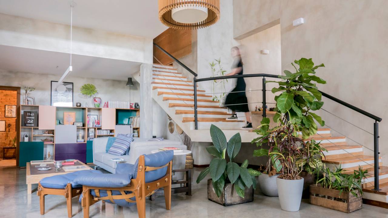 Imagem mostra sala de estar com móveis coloridos, como a poltrona mole de Sérgio Rodrigues, além de uma escada para o piso superior e diversos vasos com plantas tropicais.
