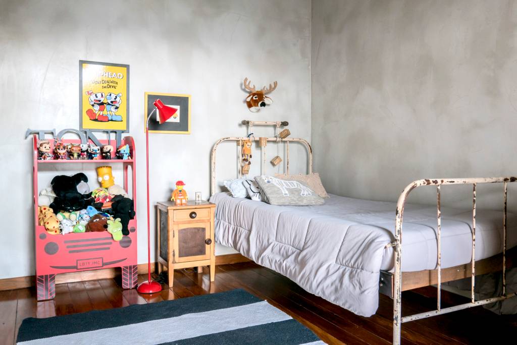 reforma-casa-veraneio-vira-endereço-principal-filipe-augusto-quarto-infantil-cama-restaurada-upcycling imagem mostra cama de ferro com cara de bem antiga, mesinha de apoio com gaveta e alguns brinquedos, quadros, luminária de chão na cor vermelha e tapete listrado sobre piso de madeira.