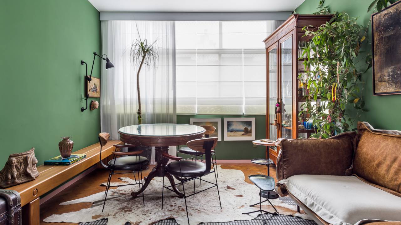 Sala de estar com paredes verdes; mesa de jantar redonda; tapetes; sofá em couro; banco em madeira