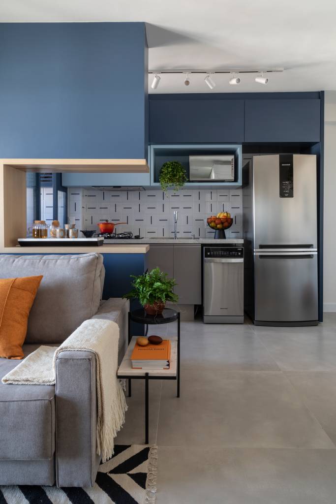 Confira-dicas-para-aqueles-que-pretendem-morar-sozinhos-Evelyn-Muller-ambientes-móveis-decoração-espaços-integrados-cozinha Vista da sala de estar e cozinha integradas. Em primeiro plano, vê-se o braço do sofá cinza, decorado com almofadas e uma manta. Ao fundo, a cozinha de móveis embutidos.