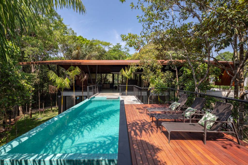 casa-na-floresta-conforto-termico-menos-impactos-ambientais-joana-frança-piscina-pavimento-superior-area-estar-aberta-paisagismo Foto mostra piscina e deck com espreguiçadeiras em primeiro plano, com varanda coberta ao fundo, com estar e jantar. Tudo no piso superior.