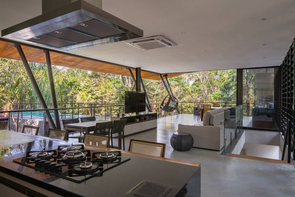 casa-na-floresta-conforto-termico-menos-impactos-ambientais-joana-frança-varanda-coberta-estar-jantar-piscina-pavimento-superior Foto mostra parte da bancada da cozinha e da sala de jantar e estar, integradas à paisagem graças às grandes aberturas de vidro, de correr.