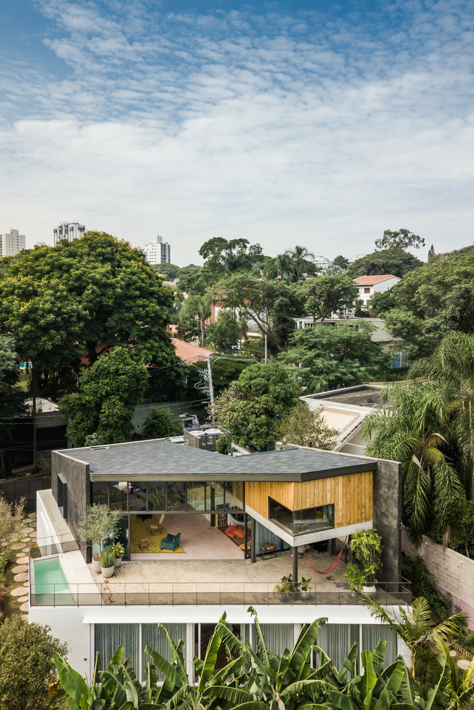 casa-sp-área-social-piso-superior-joão-morgado-vista-aérea-fachada-varanda Vista aérea mostra casa com grande varanda e sala de estar no piso superior, em meio à paisagem urbana com bastante vegetação ao redor.