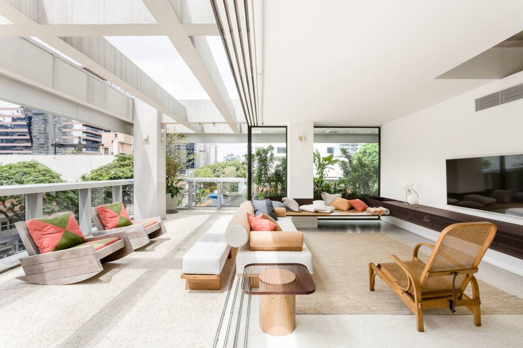 Sala de estar em apartamento tem varanda integrada e coberta com pérgula de vidro. Ambiente claro, com luz natural e móveis de madeira com almofadas coloridas.