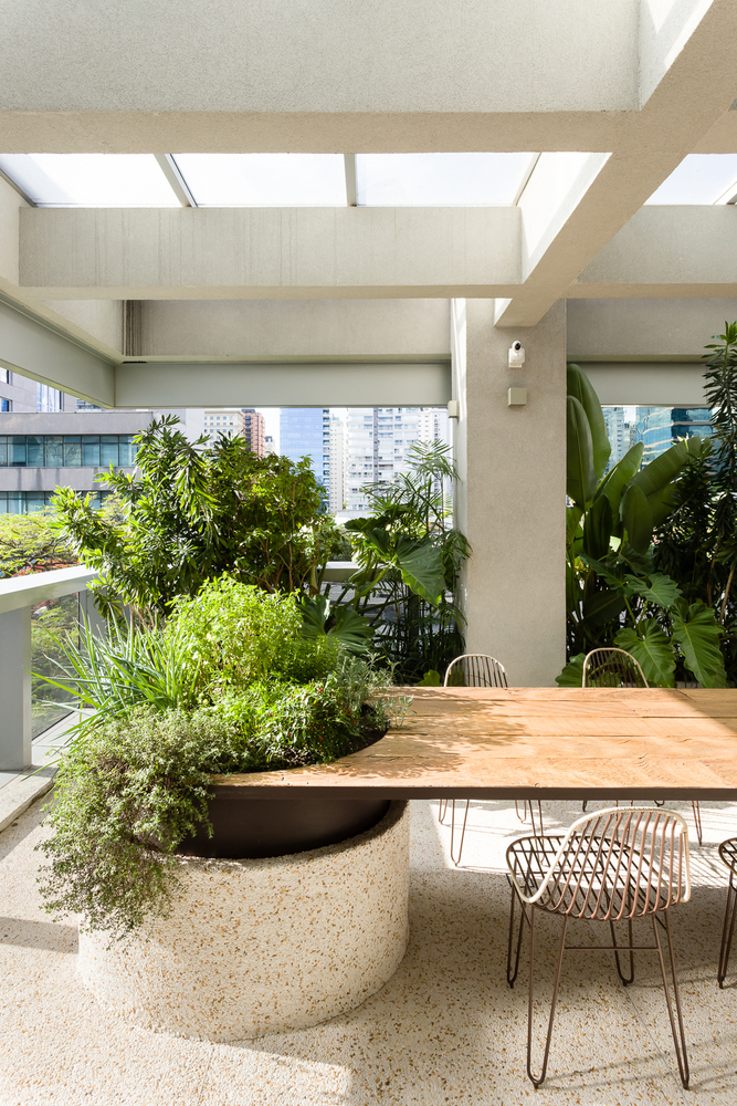 Apartamento Cel. Joaquim Ferreira Lôbo Mesa de jantar com um vaso grande de ervas e especiarias que afloram do tampo da mesa em madeira. A mesa está sob um pergolado de concreto e vidro, com um jardim tropical ao fundo.