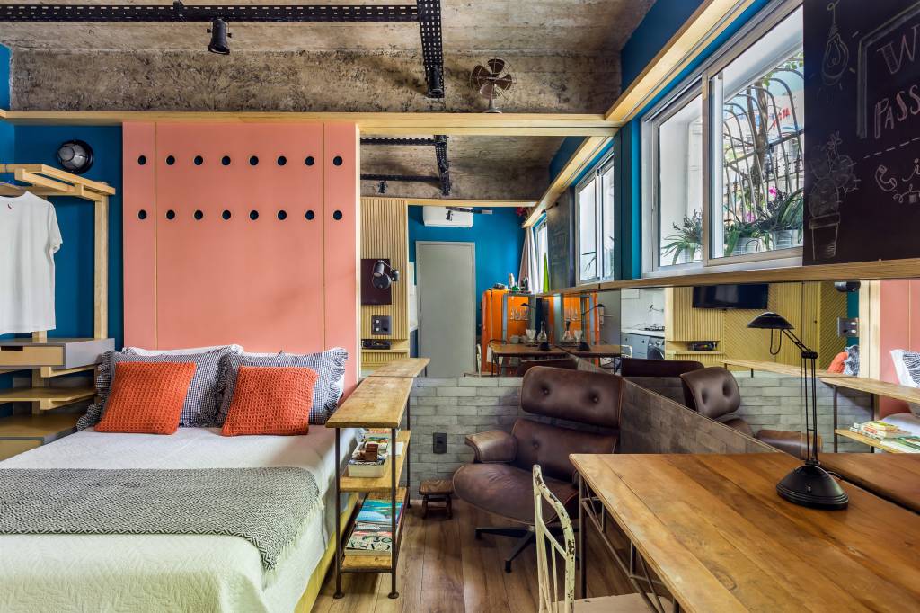 1-micro-loft-ganha-tijolinhos-e-geladeira-retro-laranja Mini loft integrado com mesa de madeira, cama e painel rosa, poltrona eames