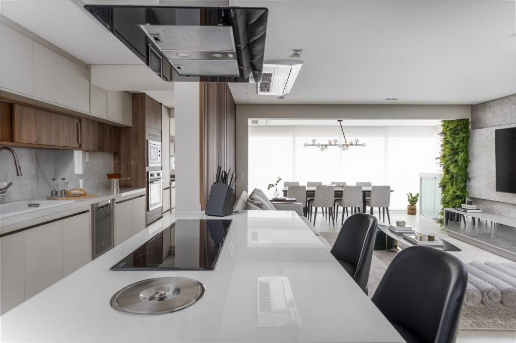 Conheça-principais-opções-bancadas-cozinha-banheiro-área-gourmet-JP-Image-bancada-porcelanato-cooktop-coifa-cozinha-integrada Foto mostra bancada de cozinha em revestimento branco, com cooktop, duas cadeiras de refeições e coifa. Ao fundo vemos a sala de jantar e uma varanda.