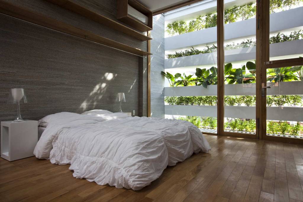 Foto mostra quarto de casal com cama sobre piso de madeira e parede da cabeceira em pedra natural na cor bege. A parede lateral é coberta por caixas de concreto com muitas plantas, que formam um jardim vertical que permite entrada de luz natural.