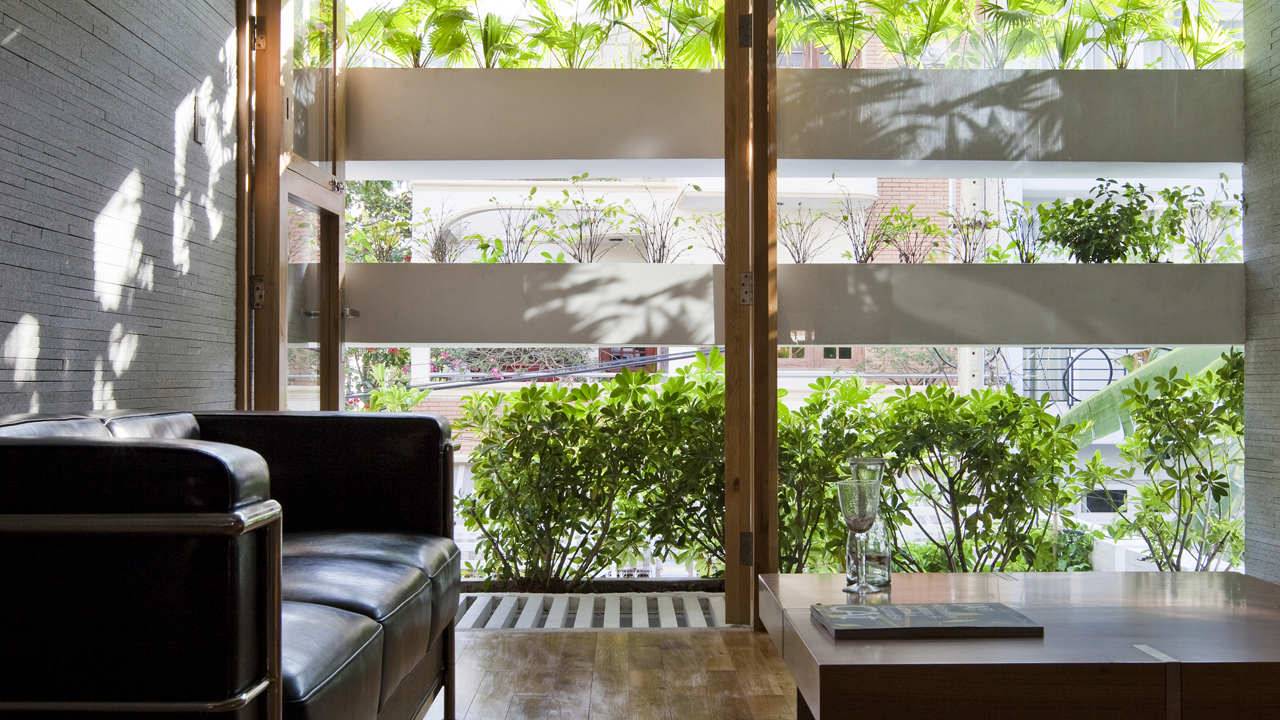 Foto mostra sala de estar com sofá preto de couro e mesa de centro em primeiro plano, com parede externa transparente, composta por caixas de concreto que recebem plantas, formando um grande jardim vertical que permite a entrada de luz natural ao ambiente.