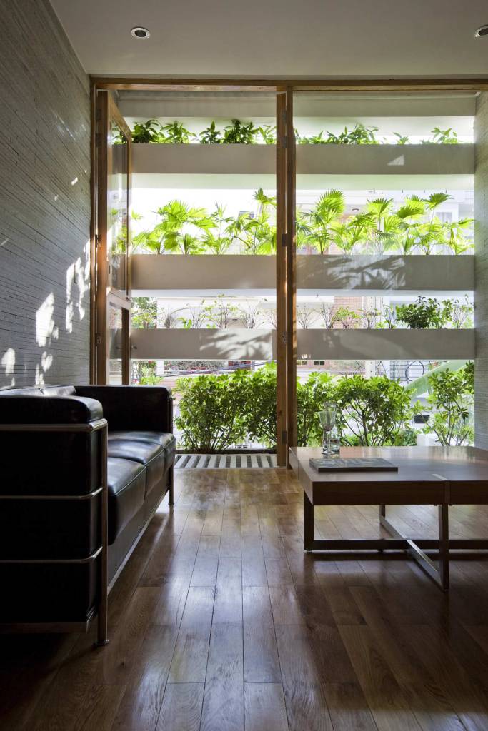 Foto mostra sala de estar com sofá preto de couro e mesa de centro em primeiro plano, com parede externa transparente, composta por caixas de concreto que recebem plantas, formando um grande jardim vertical que permite a entrada de luz natural ao ambiente.