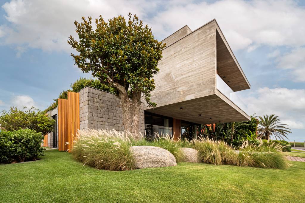 casa-litoral-gaucho-brutalismo-e-elegancia-da-madeira-Cláudio-Fonseca-ArchImage-fachada-concreto-em-balanço-parede-pedra-paisagismo-capim Foto mostra fachada de casa em concreto aparente com aberturas em madeira. Há um piso superior em balanço e jardim com gramado ao redor.