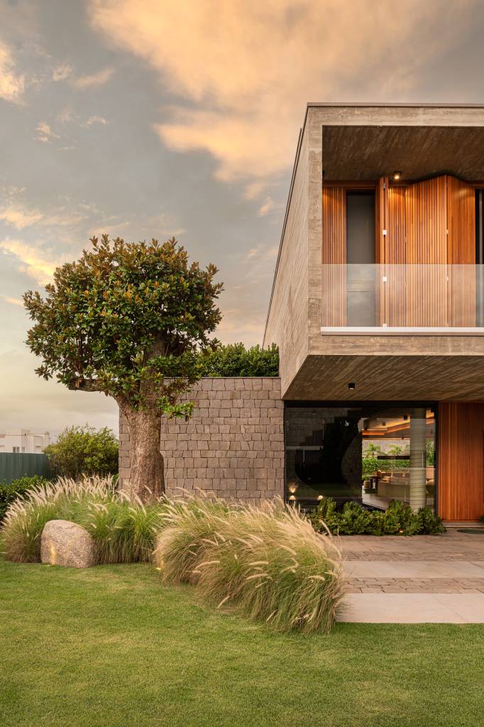casa-litoral-gaucho-brutalismo-e-elegancia-da-madeira-Cláudio-Fonseca-ArchImage-fachada-concreto-pedra-madeira-paisagismo-elegante Foto mostra fachada frontal da casa, com muro de pedra, jardim com árvore grande e casa com concreto aparente e portas e janelas em madeira.