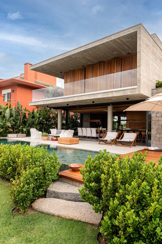 casa-litoral-gaucho-brutalismo-e-elegancia-da-madeira-Cláudio-Fonseca-ArchImage-fachada-posterior-piscina-varanda-externa-integrada-cozinha-gourmet Foto mostra casa com piscina retangular, varanda com cadeiras integrada à área de estar e o piso superior em ripado de madeira e concreto aparente.