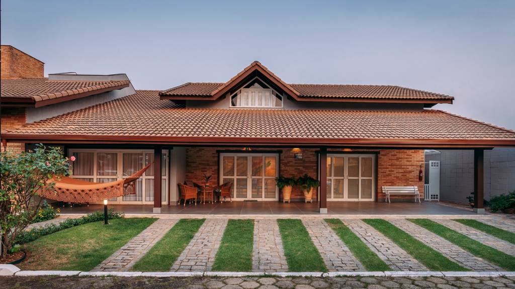 fachadas-como-ter-projeto-pratico-seguro-e-marcante-henrique-ribeiro-fachada-estilo-rustico-tijolinhos-rede-varanda-telhado-ceramico Fachada em estilo rústico com grande varanda e três pontos de aberturas (portas de correr), telhado de telhas cerâmicas e, acima, uma espécie de sótão ou mezanino com janelinha triangular. à frente, calçada que intercala faixas de grama e calçamento com pedras.