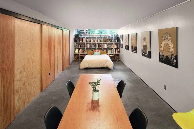 8-maneiras-renovar-piso-sem-quebra-quebra-Shed-Architecture-Design-sala-estar-piso-cimento-queimado Foto mostra mesa de madeira com 4 cadeiras pretas em ambiente com piso de cimento queimado e, ao fundo, uma cama de casal e estante de livros revestindo toda a parede.