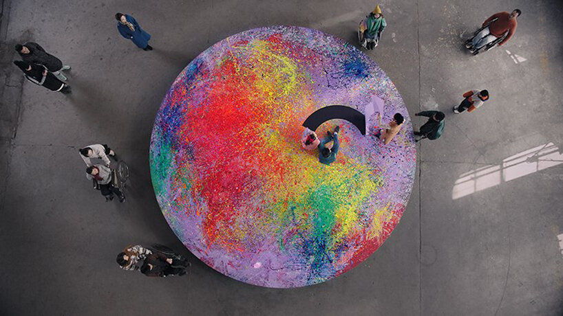 Esta instalação foi criada por pessoas com deficiência com a mente Designboom (7) Plataforma circular coberta por várias cores de tinta. Duas pessoas estão sobre ela fazendo grandes traços pretos.