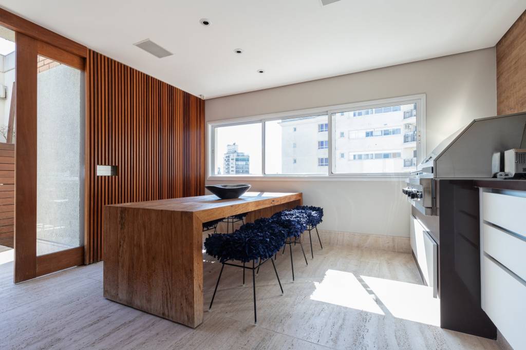 como-escolher-portas-e-rodapes-Maura-Mello-cozinha-mesa-madeira-banquetas-azuis-painel-ripado-porta-correr Foto mostra cozinha com mesa de madeira e banquetas em tecido azul marinho. Na lateral esquerda vê-se um painel ripado de madeira que 'esconde' a porta de correr, também de madeira.