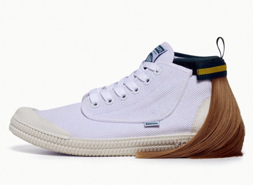 isso é um sapato com um mullet designboom volley (3) Tênis todo branco com uma mecha de cabelo presa na parte superior traseira, simulando um mullet. O cabelo é preso por uma tira de velcro que por fora é verde com uma listra amarela.