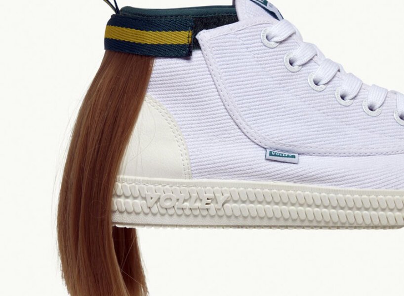 isso é um sapato com um mullet designboom volley (4) Tênis todo branco com uma mecha de cabelo presa na parte superior traseira, simulando um mullet. O cabelo é preso por uma tira de velcro que por fora é verde com uma listra amarela.