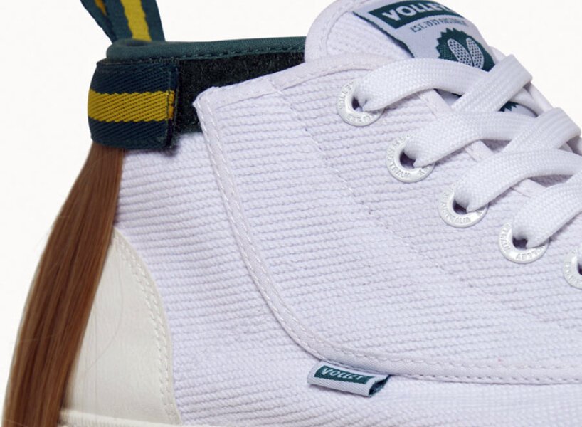 isso é um sapato com um mullet designboom volley (5) Tênis todo branco com uma mecha de cabelo presa na parte superior traseira, simulando um mullet. O cabelo é preso por uma tira de velcro que por fora é verde com uma listra amarela.