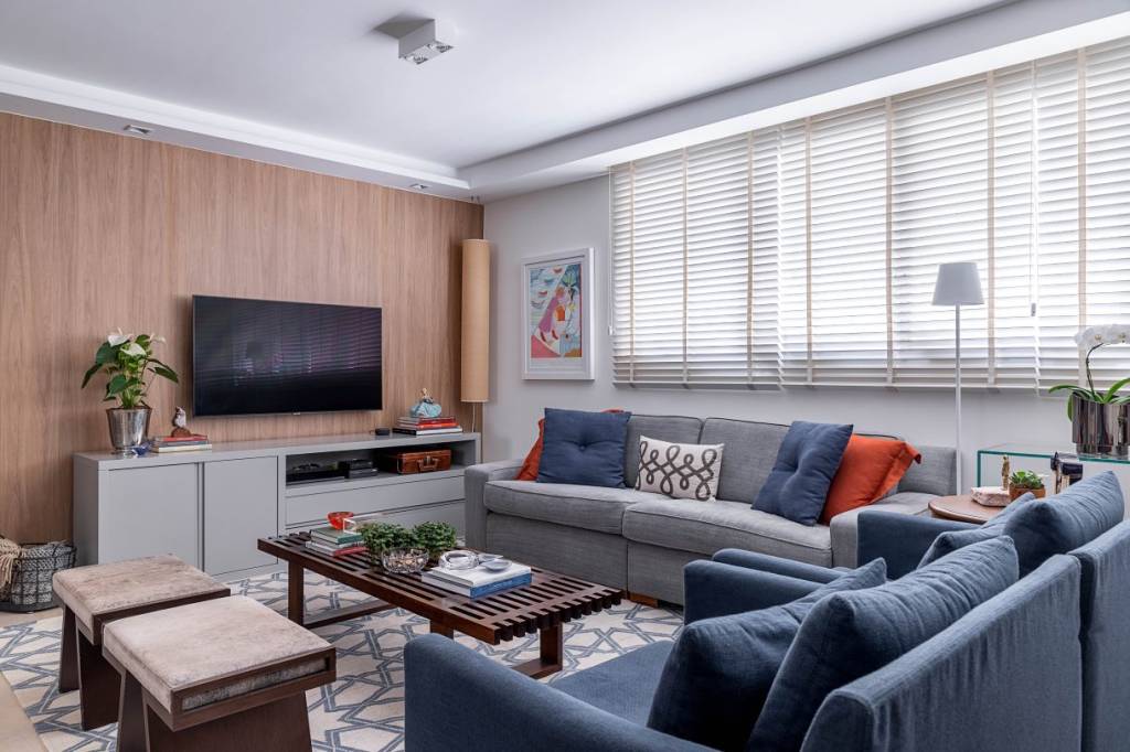 moveis-usados-nova-decoracao Visão angular da sala, dois sofás, um azul e outro cinza, grande tapete branco com detalhes geométrico em azul, televisão presa a um painel de madeira
