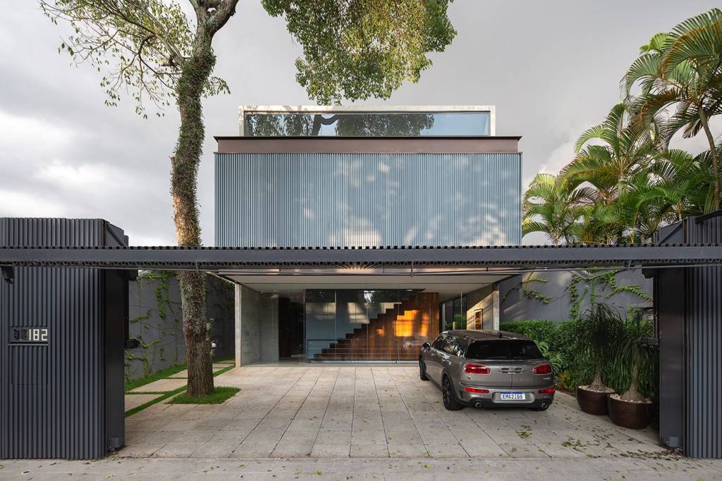 oasis-aço-madeira-concreto-andré-mortatti-fachada-frente-conteiner-discreta-integração-jardim Fachada da frente tem portão metálico da garagem levantado, onde se vê a integração do espaço do térreo com o jardim da casa.