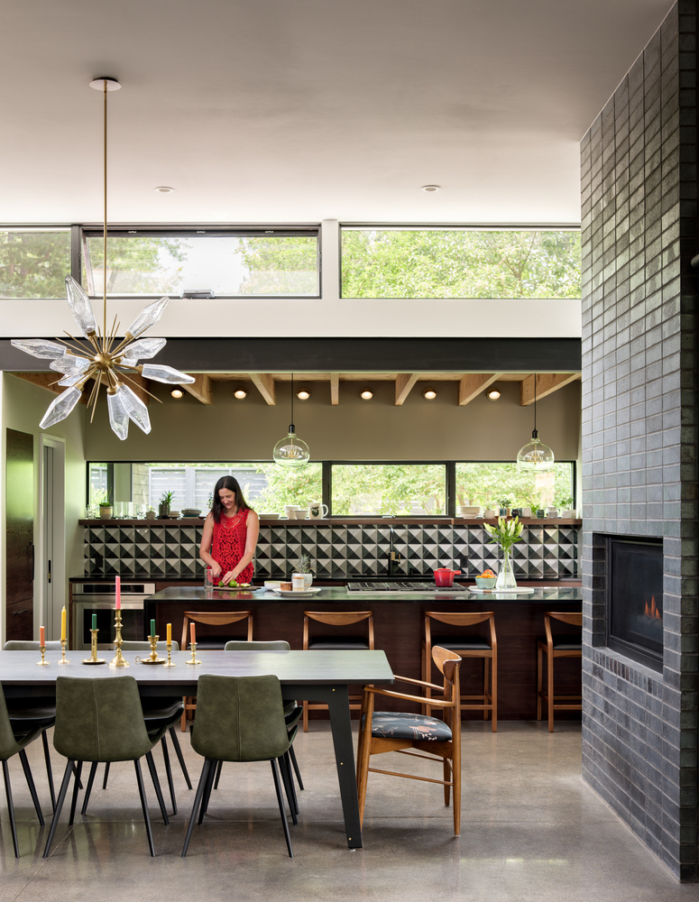 Foto mostra mulher na bancada da cozinha e à sua frente uma mesa de jantar com diversas cadeiras. Atrás dela, há aberturas com vidro no pé-direito alto, que garante luz natural ao espaço.