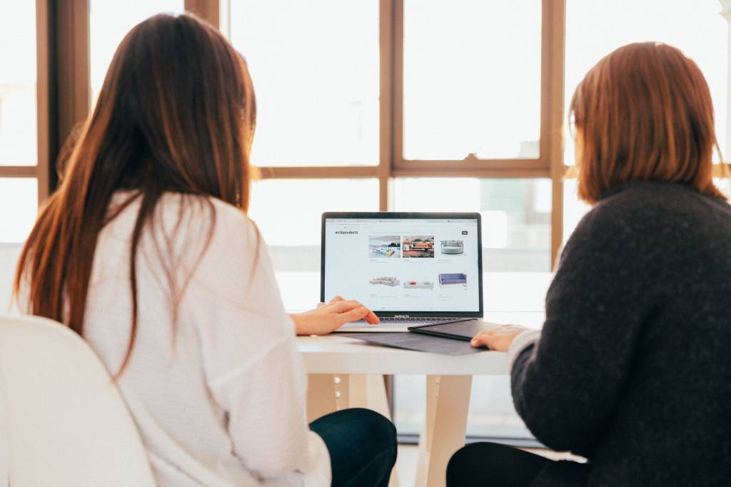 Foto mostra duas mulheres em frente a um computador observando a tela que mostra sofás e outros produtos para a casa.