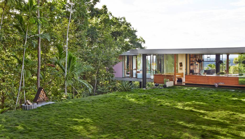 Gramado verde intenso com casa ao fundo repleta de aberturas de vidro e vegetação ao redor bem próxima.