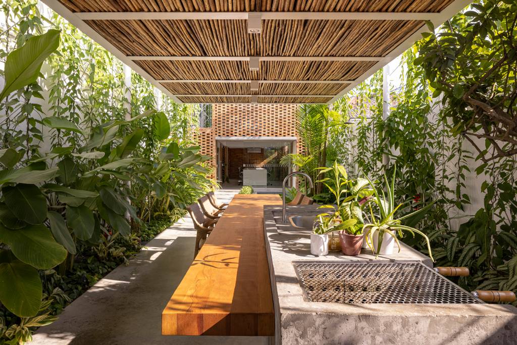 Cozinha gourmet com pergolado e aberta para o jardim tem mesa que também serve como apoio para reuniões.