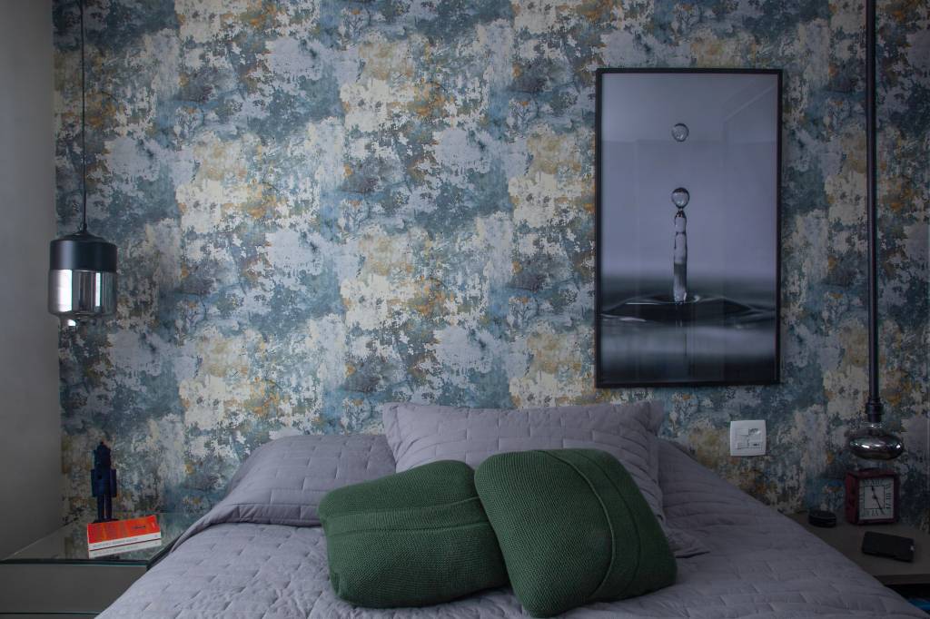 tomadas-sem-erro-Luis-Gomes-cabeceira-cama-com-tomada-papel-parede-colorido Quarto com cama de casal com colcha cinza e duas almofadas verdes e parede de cabeceira com papel de parede floral em tons de azul e bege, com quadro de gota d'água à direita e tomada ao lado da cabeceira.