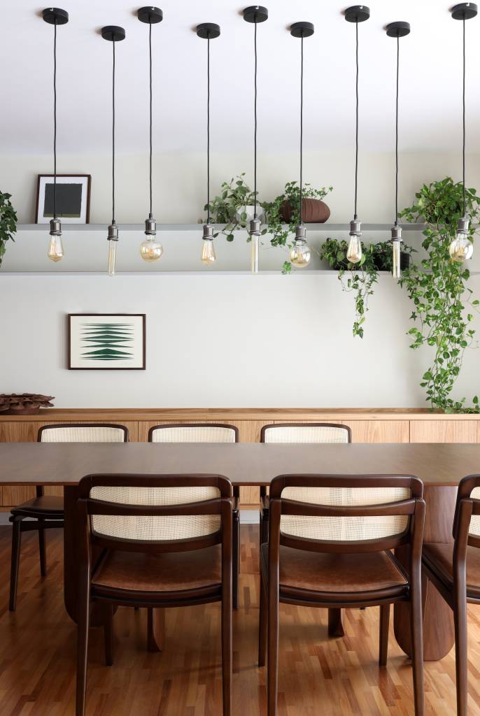 Sala de jantar com mesa de seis lugares; buffet ao fundo; luminárias pendentes e prateleiras com plantas