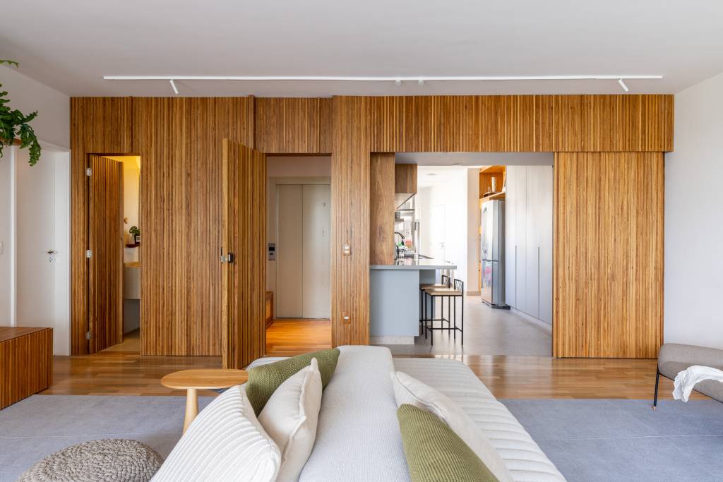 ape-integrado-140m-mesa-com-porcelanato-sofa-ilha-estudio-glik-interiores-17-porta-mimetizada Sala de estar com sofá ilha branco; painel de madeira ripada com porta de correr para cozinha