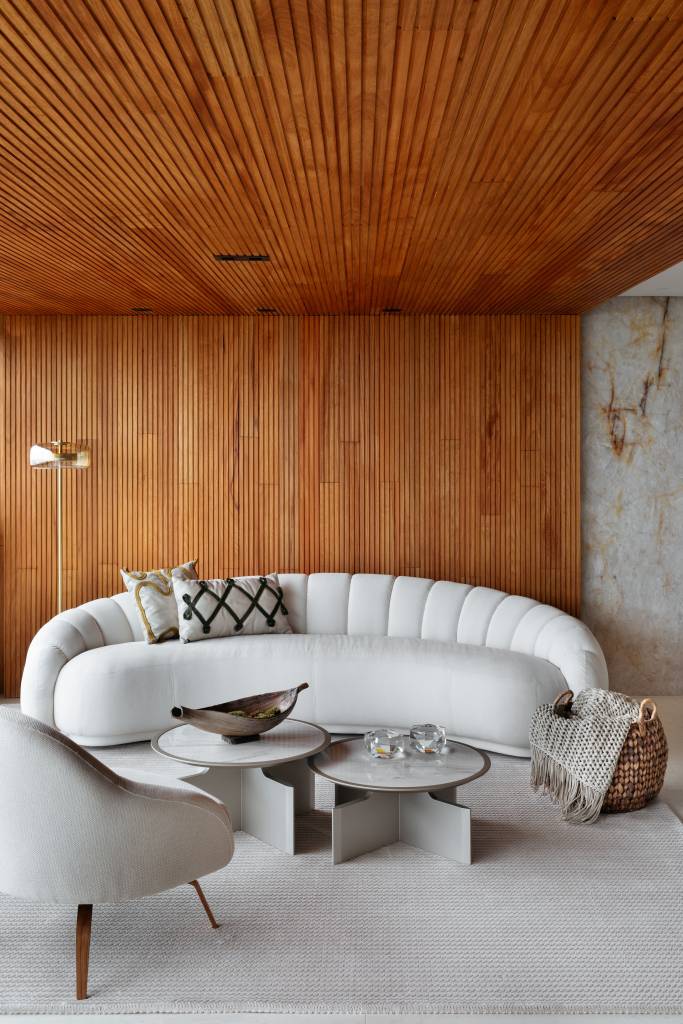 integracao-luz-natural-vista-ape-255m-paula-costa-05-sofa-curvo Sala de estar integrada com varanda; parede revestida de madeira e sofá curvo branco.