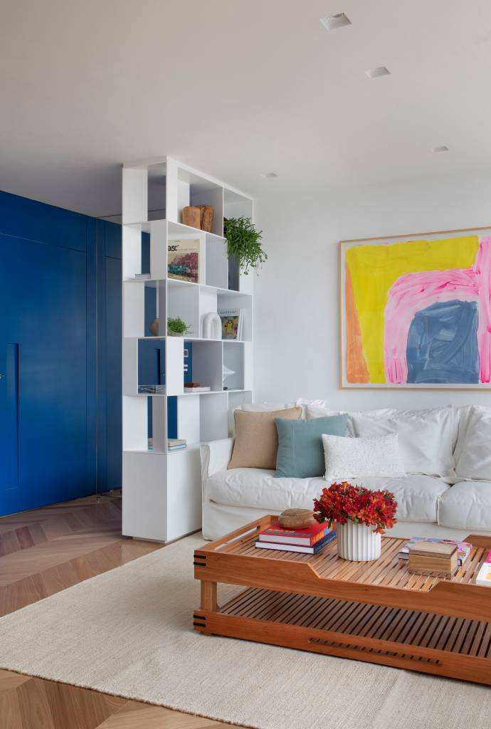 cobertura-260m-gramado-natural-escala-arquitetura-20-sala-estante Sala de estar com sofá branco, tapete bege, poltrona e quadro colorido, parede azul e estante branca vazada.