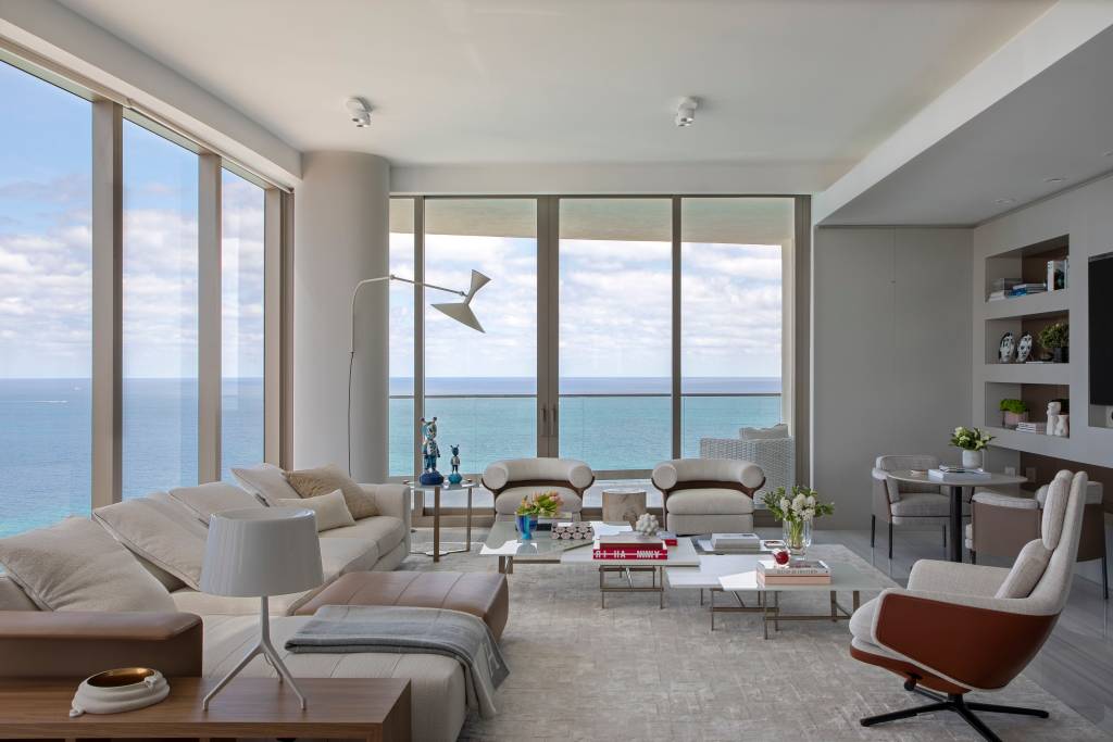 estilo-comfy-ape-450m-persiana-laminas-tecidos-daniela-saliba-02 Sala de estar branca com vista para o mar, sofá e poltrona branca, luminária branca de piso e mesa de centro.