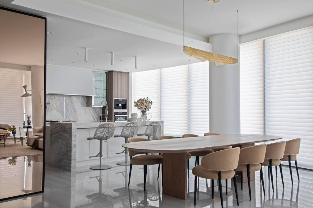 estilo-comfy-ape-450m-persiana-laminas-tecidos-daniela-saliba-13 Cozinha integrada com ilha e bancada com revestimento marmorizado e marcenaria branca. Sala de jantar com mesa de madeira.