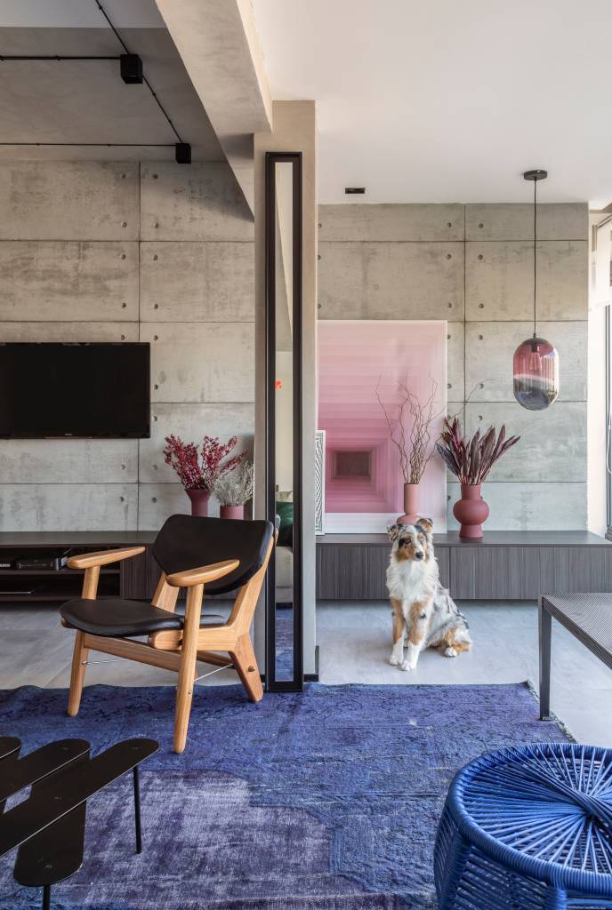 Sala de estar com paredes revestidas de cimento queimado, tapete azul, sofá cinza e poltrona rosa.