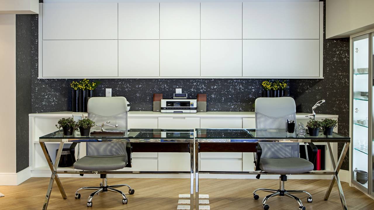 Home office para duas pessoas com marcenaria branca e mesa com tampo de vidro.