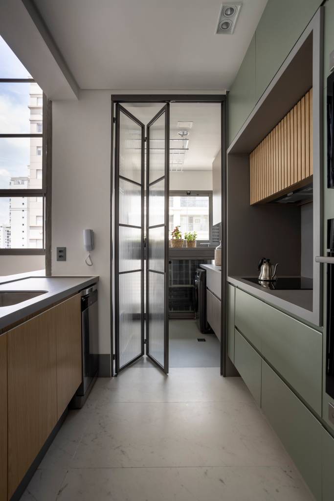 Apê de 110 m² com pé-direito duplo vira duplex com mezanino metálico Apê de 110 m² com pé-direito duplo vira duplex com mezanino metálico. NA foto cozinha separada da lavanderia por uma porta camarao de vidro canelado.