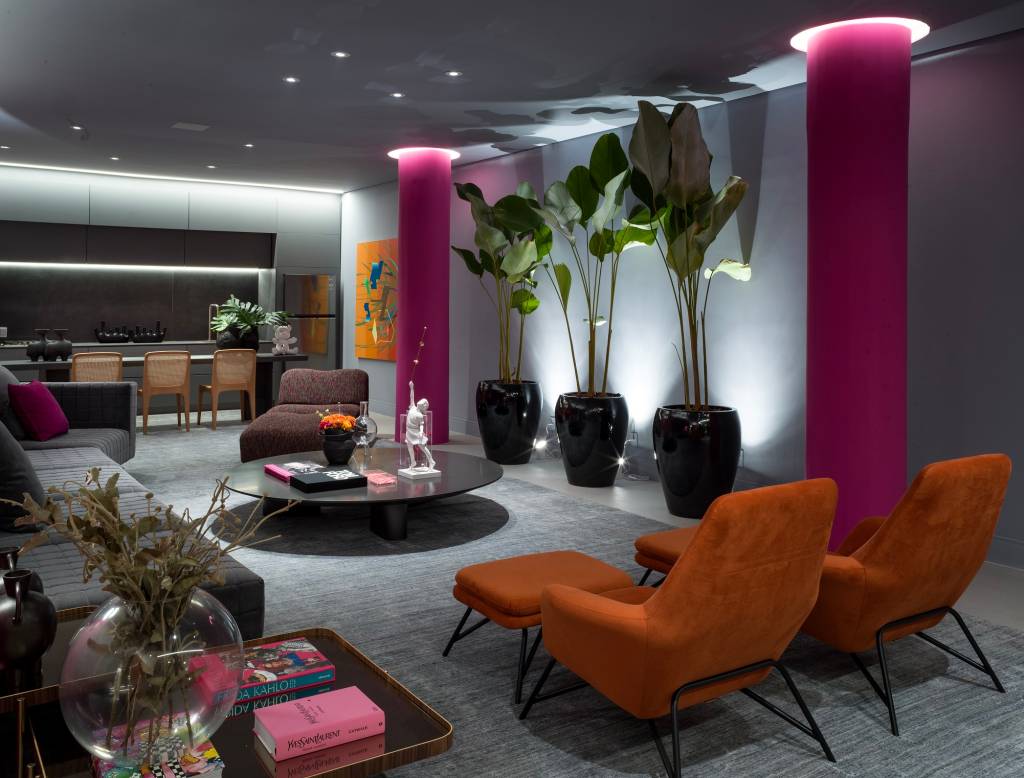 metaverso-cores-vibrantes-marcam-loft-brunete-fraccaroli-casacor-sao-paulo-2023-11 Loft com tapete cinza, mesa de centro redonda, plantas grandes e poltronas laranjas.