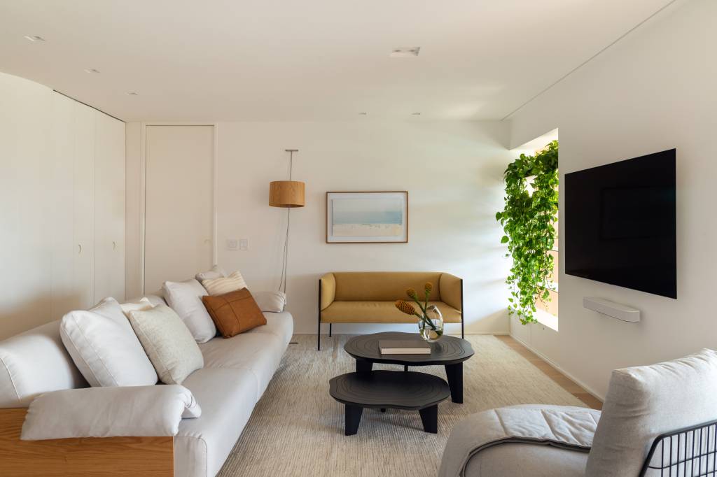 minimalista-ape-160m-varanda-integrada-formas-organicas-studio-a-g-02-sala Sala de estar com sofá branco, tapete off white, sofá em couro, mesa de centro preta.