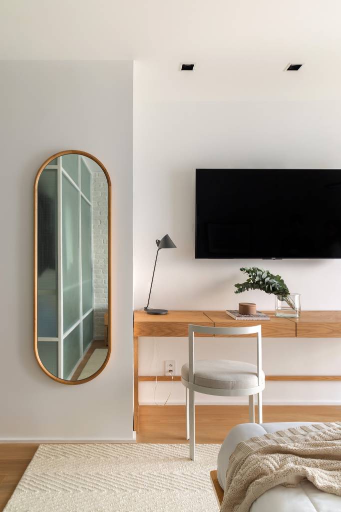 minimalista-ape-160m-varanda-integrada-formas-organicas-studio-a-g-18-quarto-branco-home-office Quarto com espelho oval e home office com bancada de madeira e cadeira branca.
