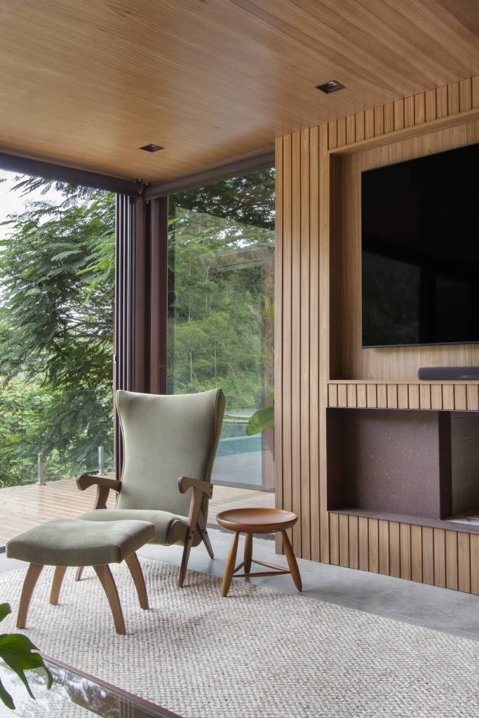 paineis-lisos-ripados-de-madeira-freijo-revestem-casa-campo-300m-diego-raposo-arquitetos-04-sala-tv Sala com portas de vidro grandes com vista para serra, parede e teto revestido de madeira, poltrona verde e painel ripado para tv.