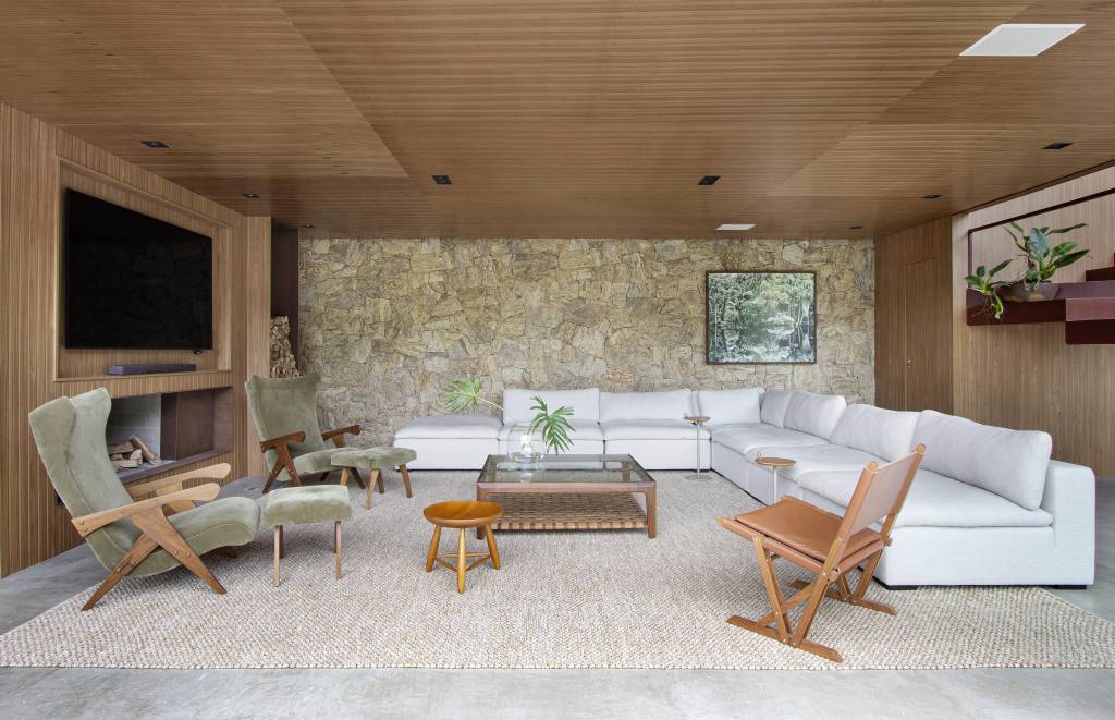 paineis-lisos-ripados-de-madeira-freijo-revestem-casa-campo-300m-diego-raposo-arquitetos-05-sala Sala de estar com parede ripada, teto de madeira e parede revestida de pedras; sala com sofá branco, tapete bege e mesa de centro de vidro.