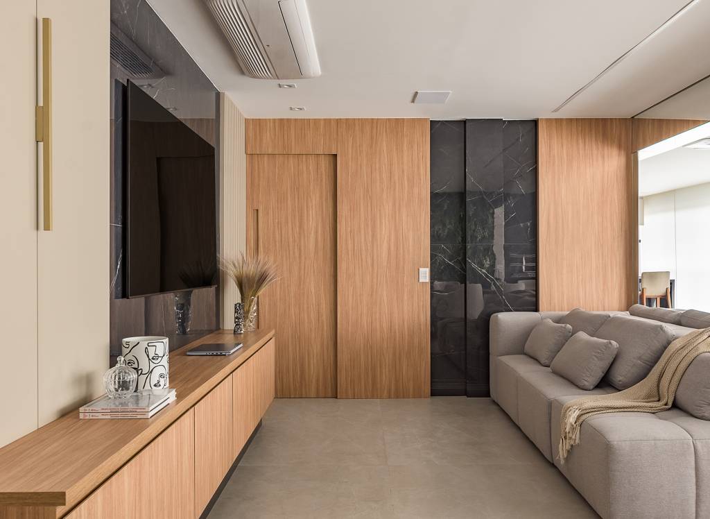 paineis-porcelanato-jardim-vertical-paredes-ape-134-m2-bianca-bahlis-credito-luiz-franco (2) Paineis de porcelanato e jardim vertical revestem paredes em apê de 134 m². Projeto de Bianca Bahlis. Na foto, sala de tv com paines de porcelanato na parede, madeira e sofá cinza.